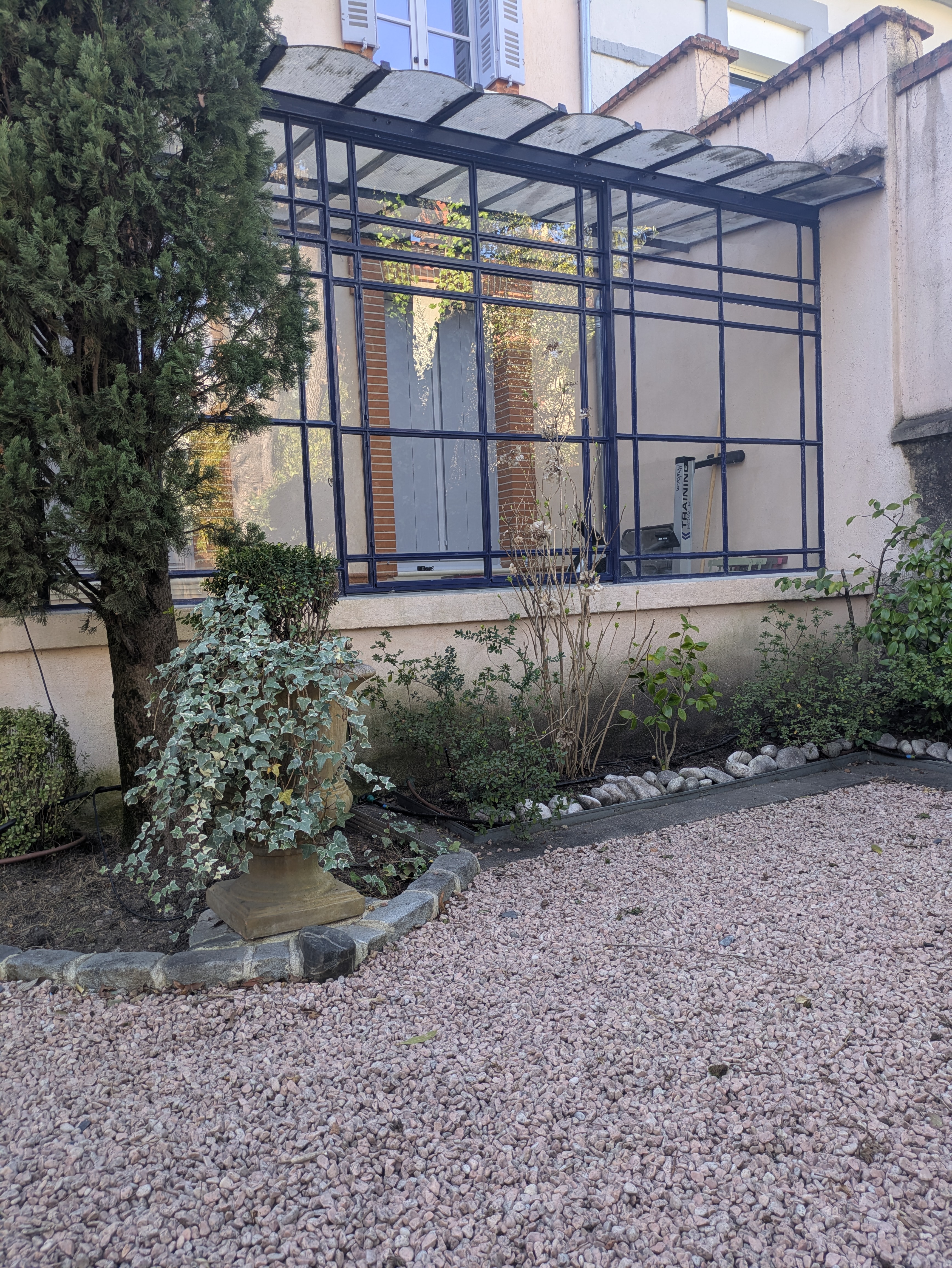 Une verrière en métal noir, une façade blanche, un jardin avec des plantes.