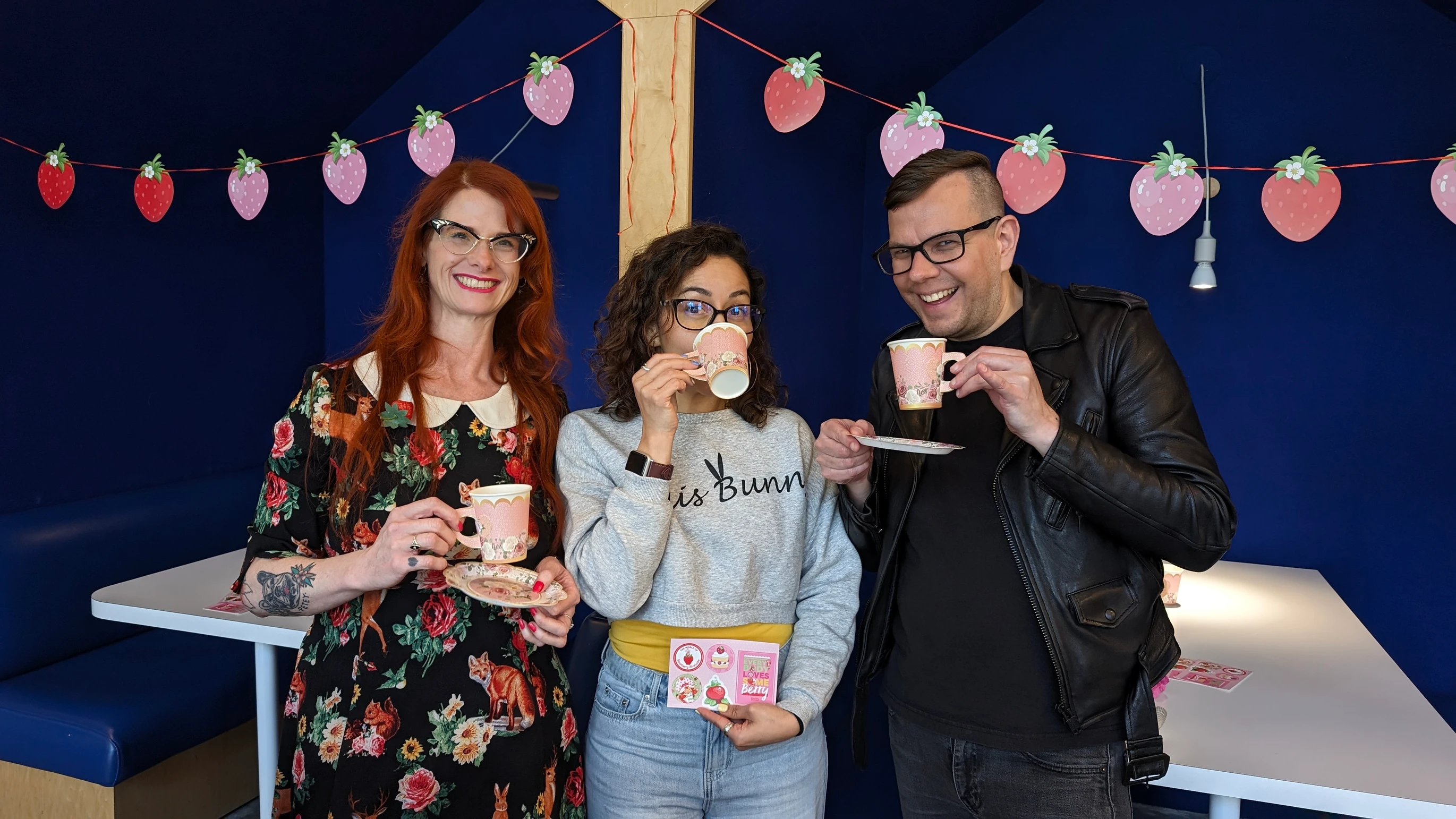 Three employees holding teacups and smiling at a decorated office table with fruit-themed bunting behind them.