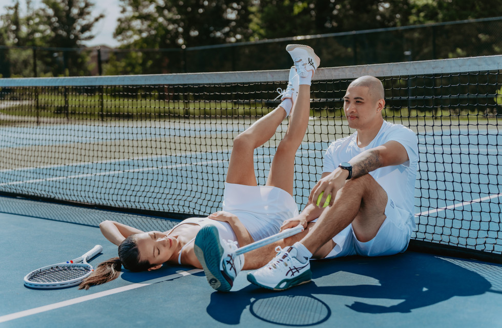 Playful shot of a couple sitting on a tennis court leaning back against the net with legs outstretched
