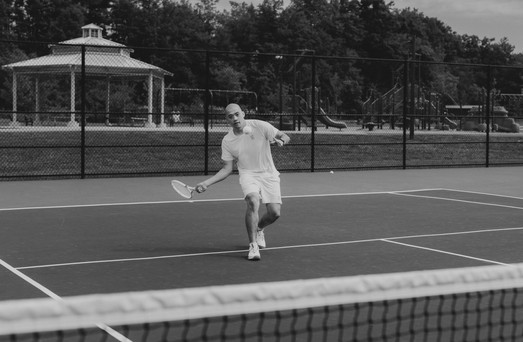 Black and white shot of a man playing tennis on an outdoor court with a gazebo in the background
