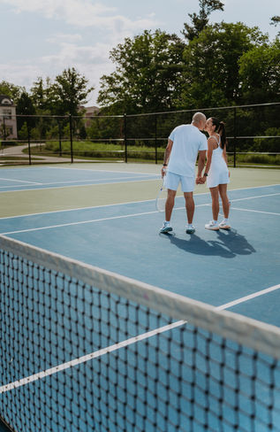 Full-length photo of a couple standing on opposite sides of a tennis court net, holding hands over the top