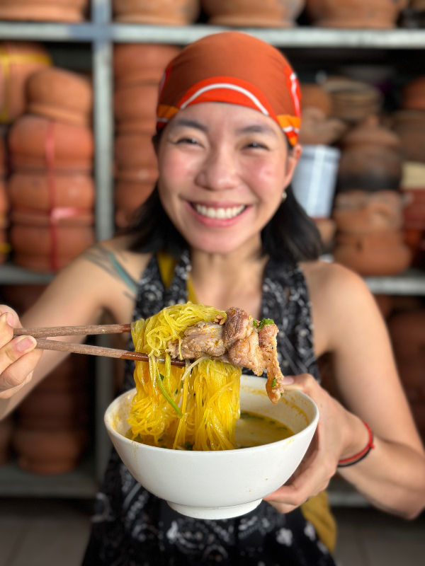 Smiling person in orange headscarf holding noodles with chopsticks over a bowl, clay pots in background. Bright and cheerful setting.