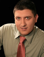 Black & white portrait of associate Dusan Gajic. Dusan has short dark hair and is wearing a medium toned shirt and tie against a dark background.