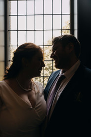 the bride and groom laugh during a wedding photoshoot next to a window