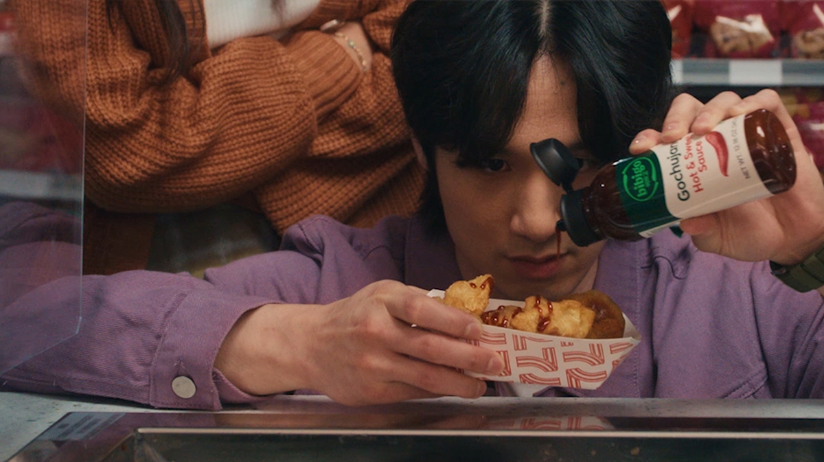 Young man in a purple shirt pours Gochujang Hot & Sweet Sauce on fried snacks at a food counter, with woman behind glass.