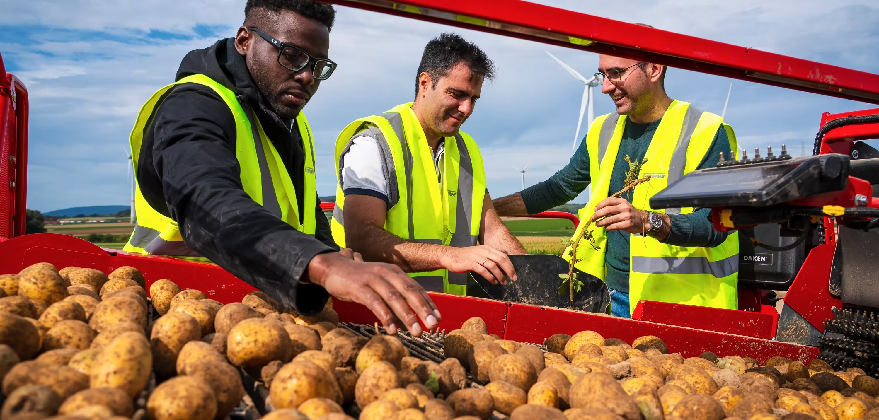 Three workers in neon vests inspect potatoes on red farm machinery in a field, with wind turbines and DAKEN label visible.