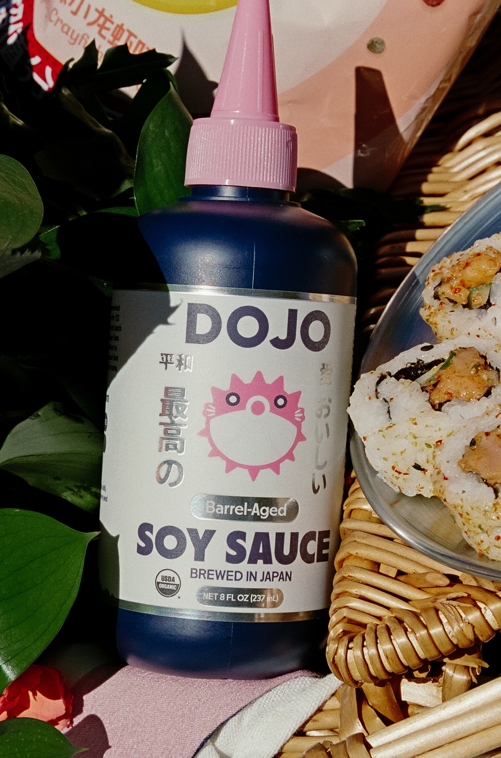 Dojo barrel-aged soy sauce bottle beside sushi in a wicker basket with green leaves and pink packaging, sunlit still life