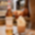 Bottle of MONIN Toasted Marshmallow syrup beside a whipped coffee mug topped with toasted marshmallows, hand adding one in a kitchen.