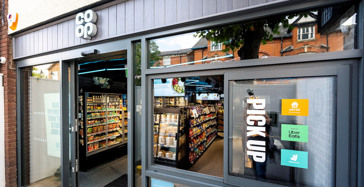 Co-op store entrance with glass doors and pickup signs for Just Eat, Uber Eats, and Deliveroo; shelves visible inside.