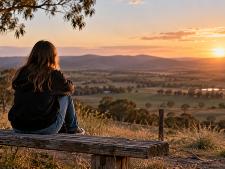 A young person sitting alone on a wooden bench overlooking the Australian countryside at sunset, reflecting a quiet moment of solitude.