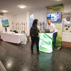 בתמונה נראה דוכן של ספק אנרגיה ירוקה נוסף בכנס בחיפה. שני נציגים משוחחים עם מבקרת ומתארים את יתרונות הפתרונות שהם מציעים. מאחוריהם שלט צבעוני עם לוגו החברה, ועל הדוכן מוצגים דגמים קטנים וקטלוגים. התאורה הנעימה מדגישה את האווירה הרצינית והירוקה של המקום