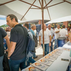 בתמונה נראית עמדת ברושורים בכנס מקצועי לאנרגיה ירוקה. מדפים או מתקן תצוגה מלאים בעלונים צבעוניים וקטלוגים, חלקם ממותגים בלוגו של חברות שונות. מבקרת עומדת ליד העמדה ובוחרת חומר לקריאה. ברקע נראים משתתפים נוספים ותאורה בהירה שמדגישה את סדר וניקיון המקום