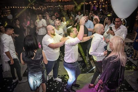 בתמונה נראים חברים לבושים בבגדים חגיגיים רוקדים במעגל או בשורה על רחבת הריקודים באולם חתונה. התאורה הצבעונית והבזקי האורות מוסיפים תחושת מסיבה, והבעות הפנים מציגות שמחה, התלהבות ורוח חברות חמה