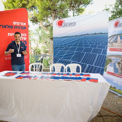 בתמונה נראה דוכן של ספק אנרגיה ירוקה בכנס, כשהרקע מעוטר בפנלים סולאריים גדולים ומוארים. נציג החברה עומד מאחורי השולחן ומסביר למבקרת על השימושים והחיסכון שבאנרגיה סולארית. על הדוכן פרוסים דגמים קטנים, טאבלט ומסמכים. התאורה הטבעית מדגישה את הנושא הסביבתי והמתקדם של התערוכה