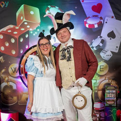 צילום של זוג עובדים מחופשים לאליסה בארץ הפלאות באירוע חברה בעיצוב קזינו. היא לובשת שמלה תכלת עם סינר לבן, והוא בדמות צבעונית מהסיפור, על רקע אורות אדומים, זהובים ושלטי קלפים. התמונה משדרת יצירתיות, שמחה ואווירה שובבה של ערב פורים קזינואי מלא קסם