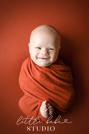 Swaddled smiling newborn boy on orange backdrop and matching orange wrap taken during newborn photos at Little Babe Studio in Middletown, Ohio