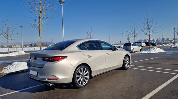 Mazda 3, light grey from the back right side on a sunny winter day on a parking with a bit of snow and naked young short trees