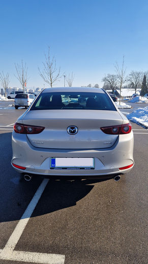Mazda 3, light grey from the back on a sunny winter day on a parking with a bit of snow and naked young short trees with the white rocky mountains in the background