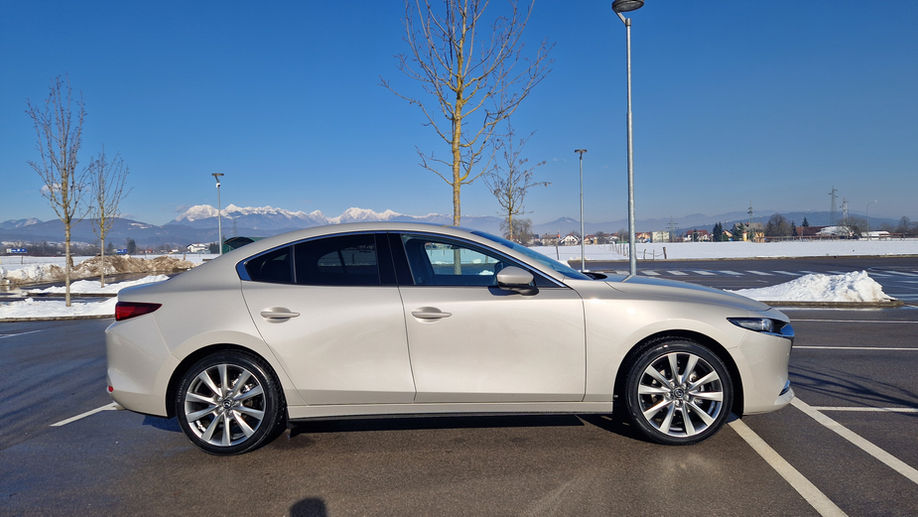 Mazda 3, light grey from the right side on a sunny winter day on a parking with a bit of snow and naked young short trees with the white rocky mountains in the background