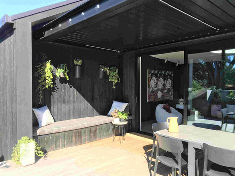 Covered outdoor patio with a built-in daybed bench, striped cushions, black timber walls, wall-mounted plants, and sliding glass doors revealing a modern interior room.