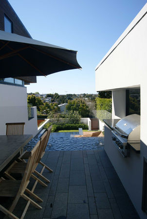 Elevated outdoor kitchen and dining patio with dark stone pavers, a built-in BBQ, and timber dining table looking down over an infinity pool in Mairangi Bay.