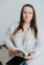 Woman, health & wellness coach, in a cozy sweater, holding an open book and a notebook on her lap, smiling slightly against a light background.