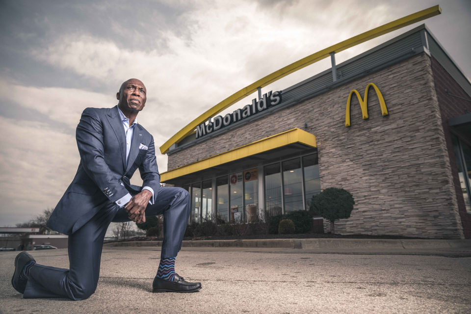 Businessman kneeling outside McDonald's under a cloudy sky, blending corporate success with grassroots storytelling.