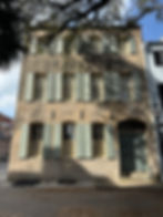 Historic beige building with pale green shutters under a tree's shade. Sunlight casts shadows on the façade. A "One Way" sign is visible.