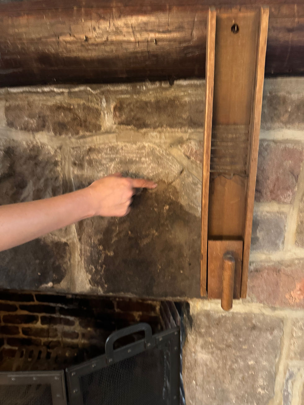 Finger pointing at a stone wall inside a rustic fireplace setting with wooden beams. A mesh fire screen is partially visible below.