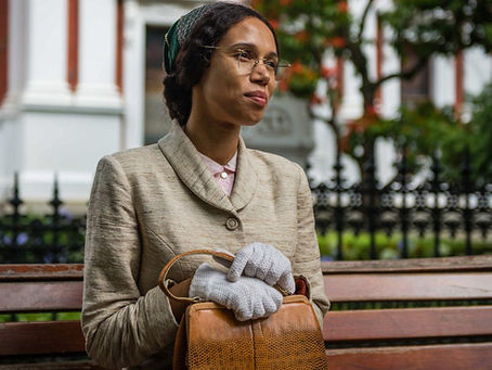 Vinette Robinson playing Rosa Parks, sat on a bench holding handbag