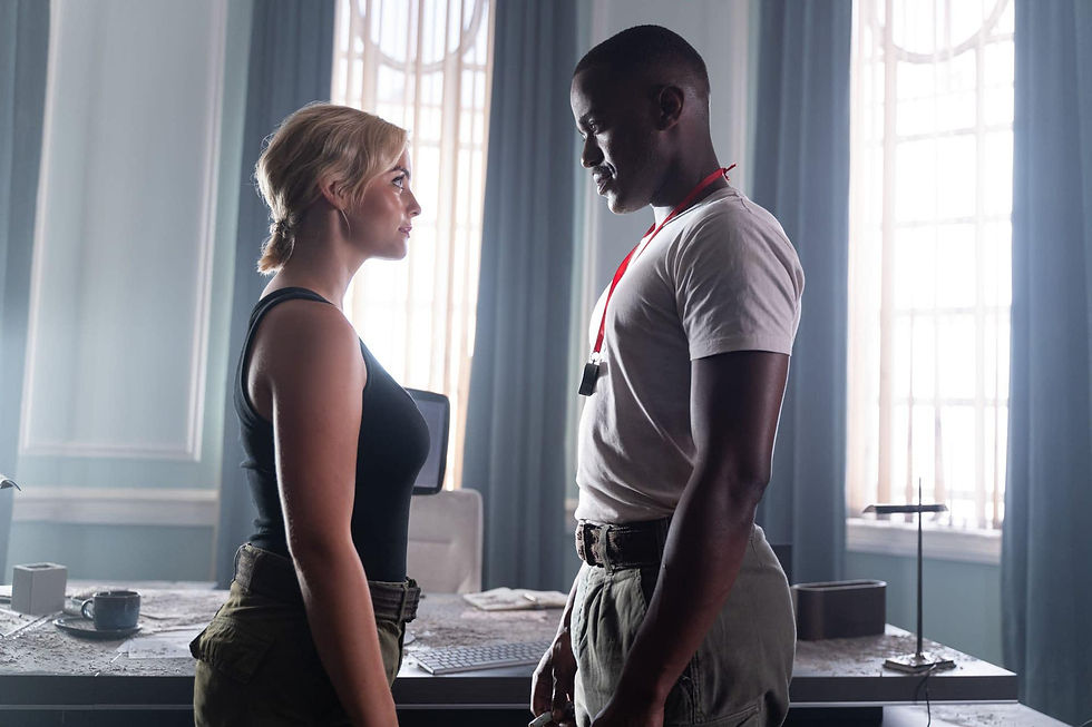 Millie and Ncuti wearing quite boring clothes by their standrads. Millie in black vest top and green trousers, Ncuti in white tshirt and trousers. He has a whistle round his neck and they are looking at each other