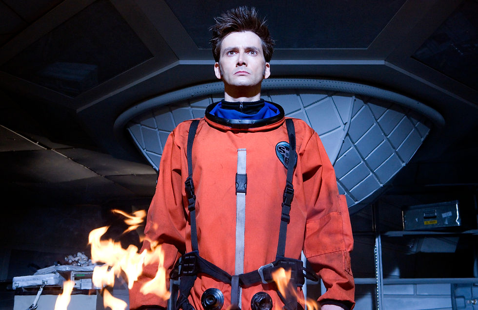 The Doctor looking focused in orange spacesuit. Fire at bottom of screen. He is about to cause some serious shit