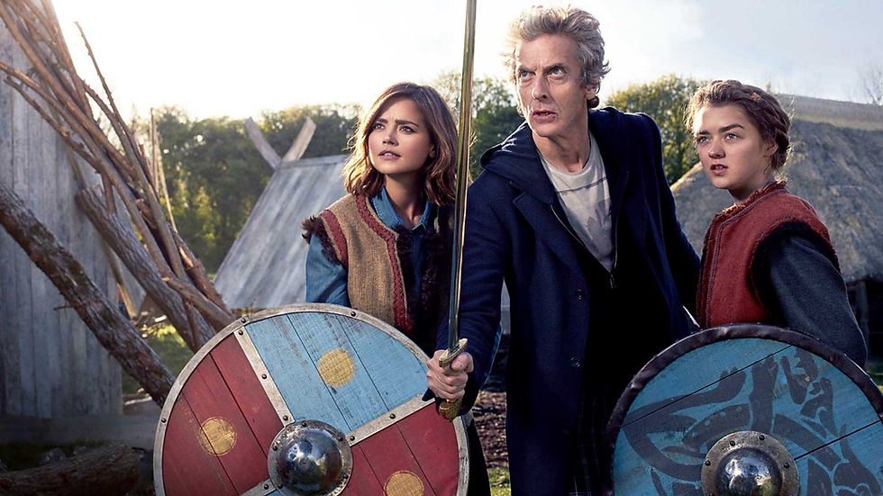 Clara and Ashildr holding viking shields with the Doctor between them holding a sword. They are in the village. Wooden houses around them.