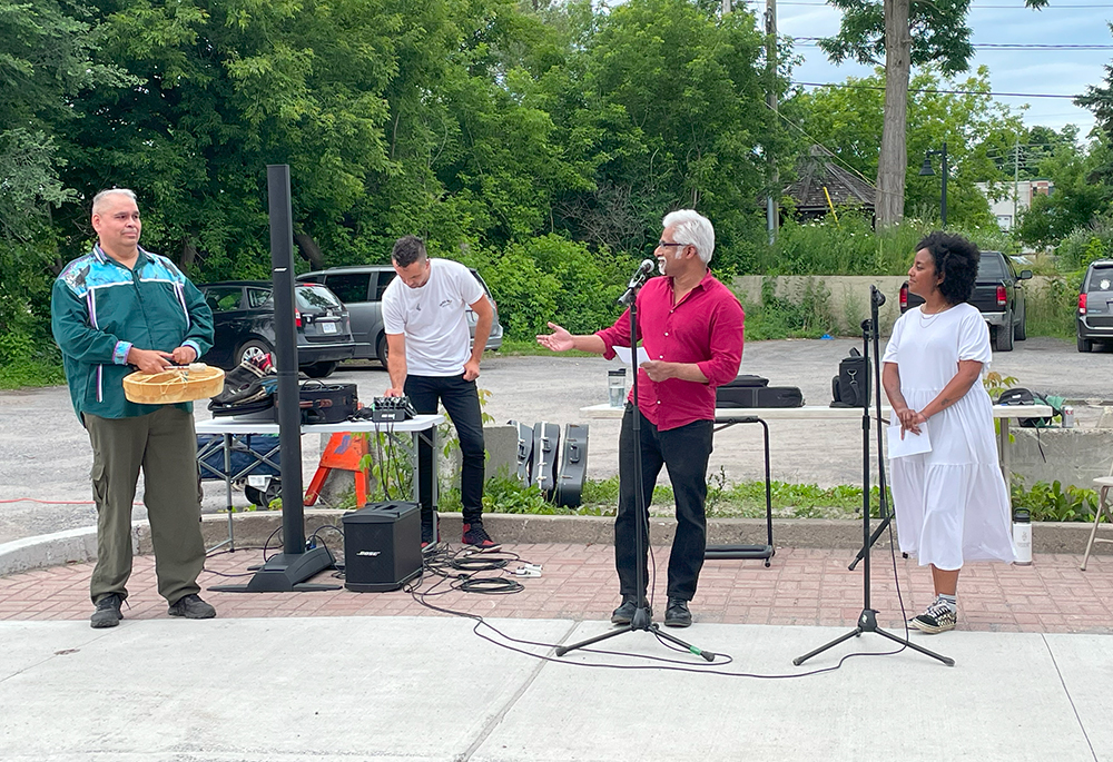 An Indian and a Black woman MCing an event, introducing an Indigenous musician about to perform at an outdoor event.