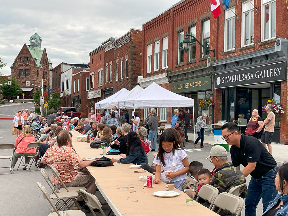 A large group of diverse people and families gathered and eating at a long table, set up outdoors on a town main street with tents and local storefronts in the background.