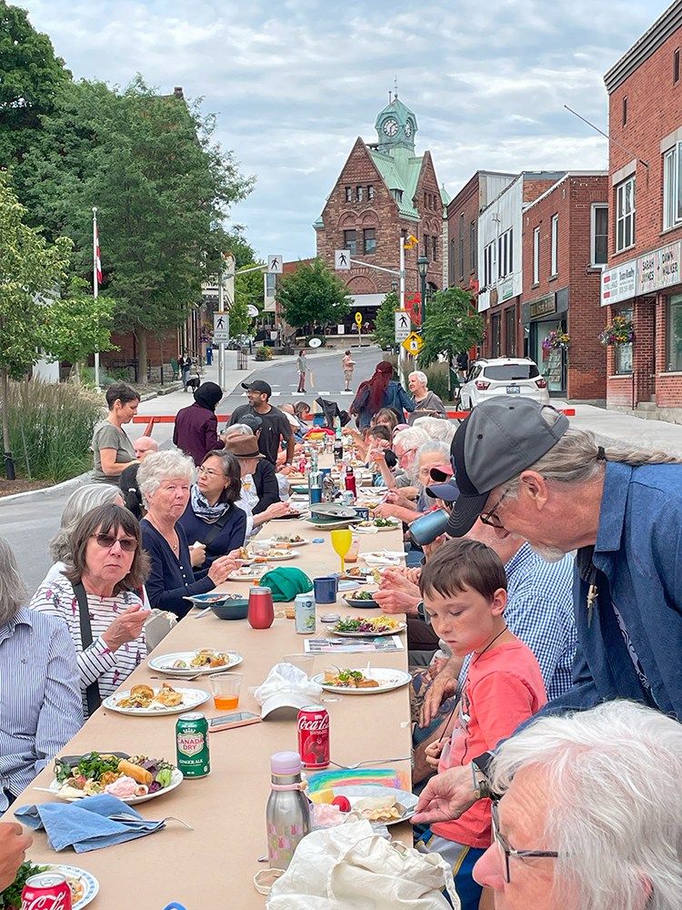 A large group of diverse people and families gathered and eating at a long table, set up outdoors on a town main street.