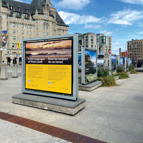 A group of outdoor exhibit panels in front of a large Victorian stone building. The front panel is bright yellow and shows an image of a yellow sunset over a beach, with the title "In the Languages of These Lands" in English and French.
