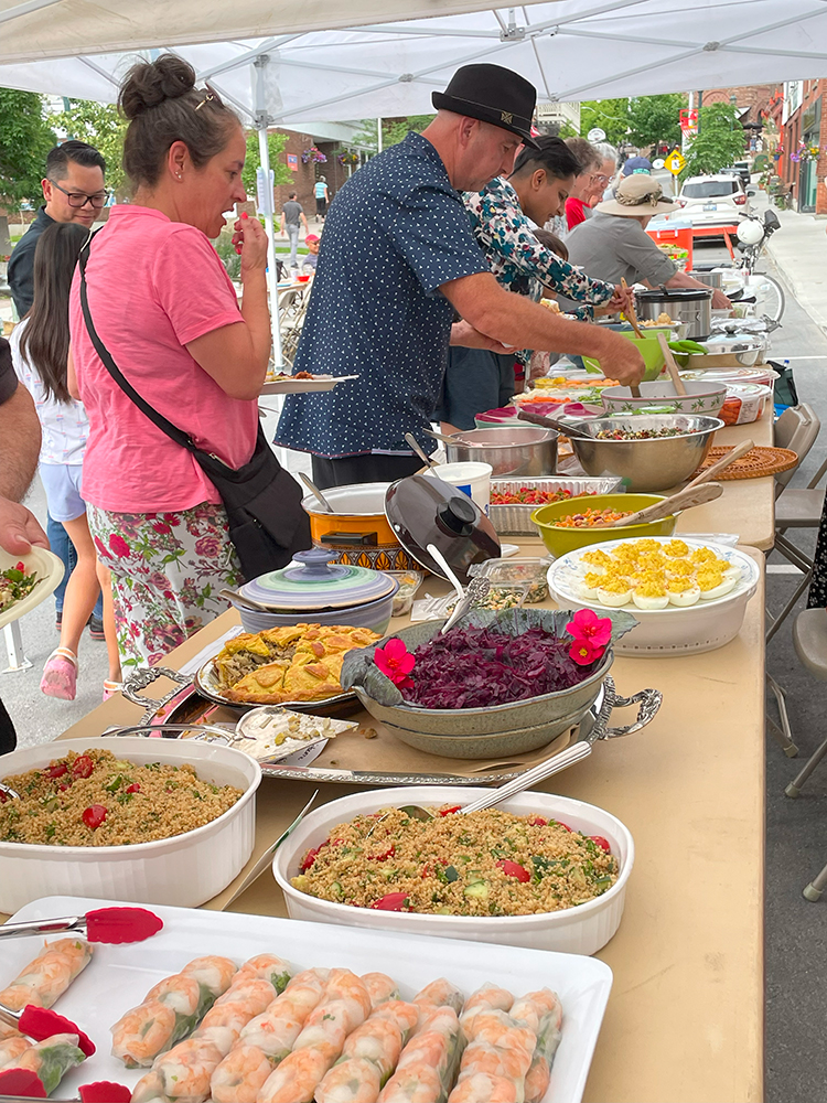 A long, tented table full of several platters and large dishes of multi-ethnic and -cultural foods, with a line of people serving themselves from each dish.