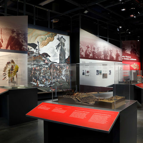 An exhibit gallery with several large panels in the background showing text, large images and a painting of First World War soldiers, with a display case containing a diorama and red text panels.