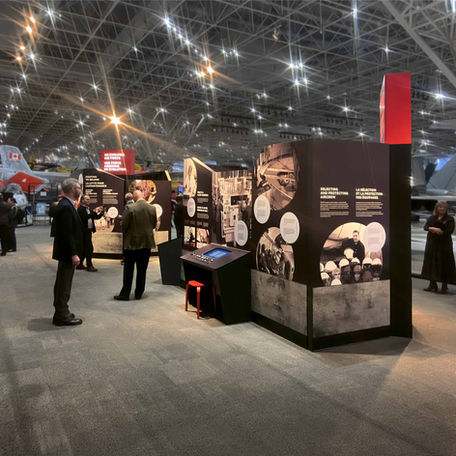 Large museum space with several full-size planes, with exhibit black and red displays with image and text content being viewed by several museum visitors