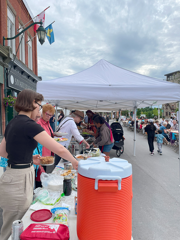 A long, tented table set up outdoors on a town street, featuring multiple plates of food, drinks, and coolers, with people gathered and serving themselves food.