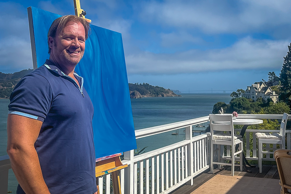 Belvedere resident Brian Higgins poses with one of his paintings on his deck Sept. 2, with the bay view that inspires much of his artwork. (Tyler Callister / The Ark)
