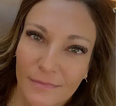 A person with long eyelashes and brown hair looks calmly at the camera. The background is neutral, creating a serene mood.