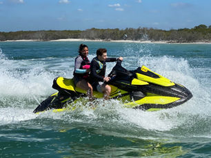 Two people ride a yellow and black jet ski in choppy water with a distant sandy shoreline. Blue sky adds a sense of adventure and fun.