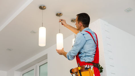 Electrician in a blue shirt and red apron adjusts hanging lights with a screwdriver in a modern, bright room. He wears a tool belt.