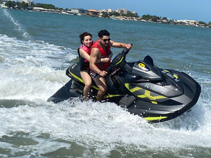 Two people on a jet ski in blue and pink vests ride on choppy water, smiling and giving a thumbs-up; green trees in the background.