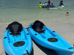 Two blue kayaks on sandy beach, people on jet skis in water, child wading. Cloudy sky, calm mood, trees in background.