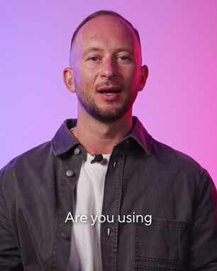 Man in white shirt gestures with hands at laptop with stickers. Pink gradient background. Appears curious or questioning.