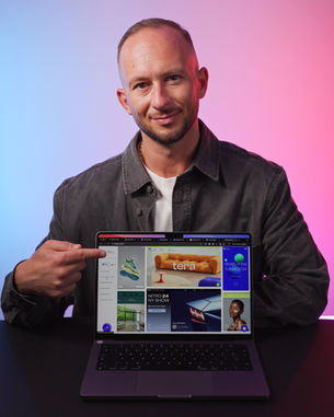 Man in a gray jacket smiles, pointing at a laptop screen with various colorful website thumbnails. Background is blue and pink gradient.