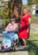 A woman in a red dress stands next to an elderly woman in a wheelchair outdoors, under a brick archway.